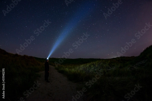 Fototapeta Adventurous man looking up at the stars on a beach path at night with his headlamp shinning up at the sky.