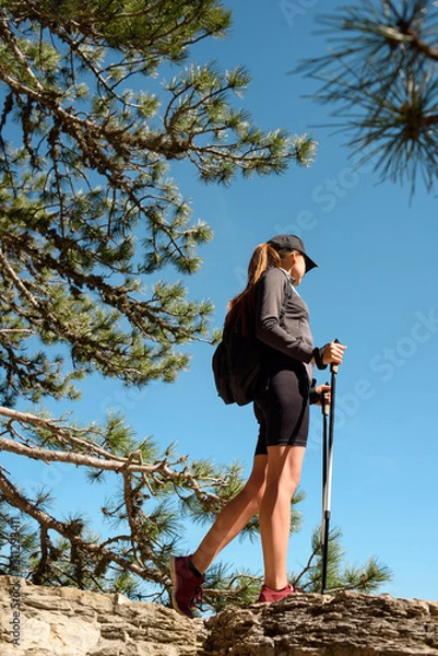 Obraz Young beautiful woman stay traveling with backpack on mountains with walking sticks on blue sky background and look away