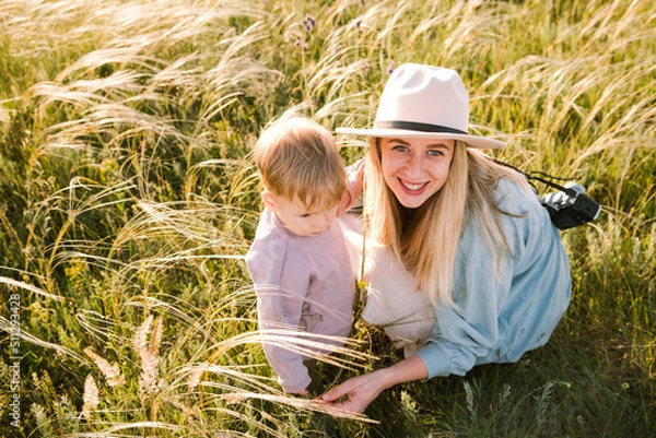 Obraz Blond happy mother in hat with camera hug her son among field, look at camera, smile