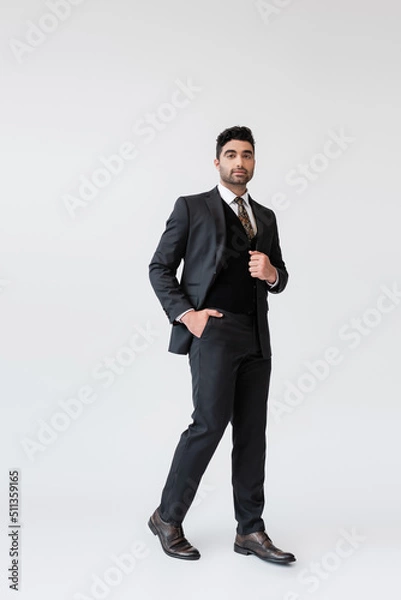 Fototapeta full length of muslim groom in formal wear standing with hand in pocket on grey.