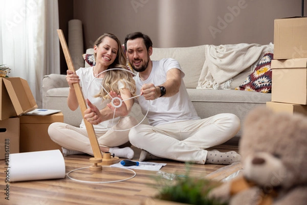 Fototapeta A happy couple in love are sitting on the floor of their new apartment, assembling living room furniture, the man holding a lampstand spinning it like a car steering wheel, fooling around.
