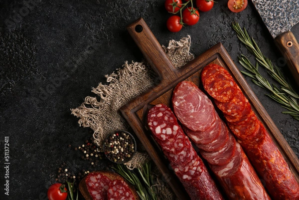 Fototapeta Sausage and cold cuts. Types of different loaf sausages on an old wooden board on black background and burlap with cherry tomatoes, rosemary, garlic and spices.Rustic.Top. Background image, copy space
