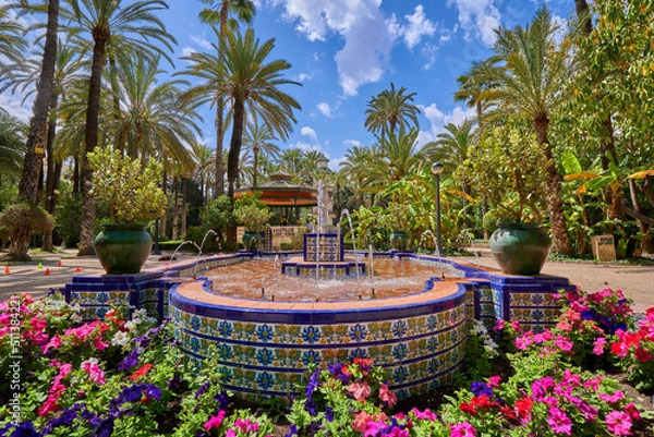 Fototapeta Fountain inside the Palmeral de Elche surrounded by flowers and palm trees. In the municipal park of Elche, Alicante, Spain