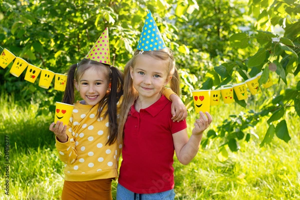 Obraz World Smile Day and Children's holiday, birthday, girlfriends in hats hugging, yellow paper cups for World emoji Day