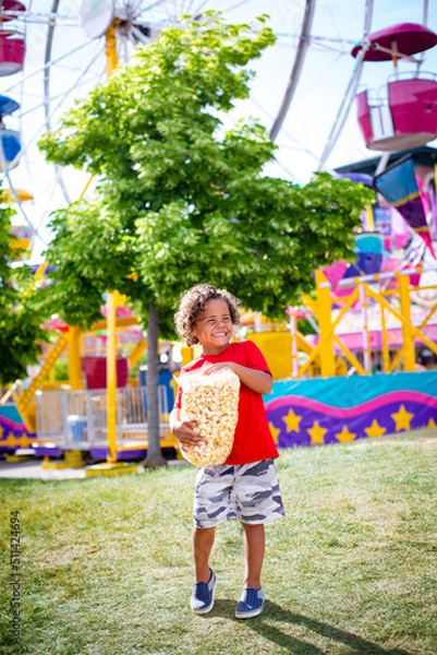 Obraz Cute bi-racial little boy eating a big bag of popcorn at an outdoor carnival. Smiling and having fun by a large ferris wheel at an amusement park