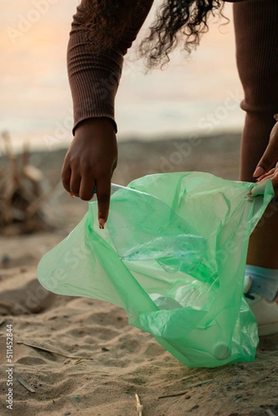 Fototapeta Unrecognizable African woman leaning down to collect trash garbage from sand on beach in green bag. Womans hand picking up plastic bottles. Ecology, environmental conservation, pollution. Vertical.