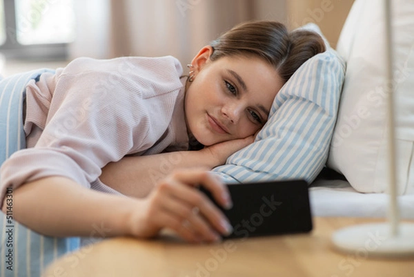 Fototapeta people, bedtime and rest concept - happy smiling teenage girl with smartphone lying in bed at home