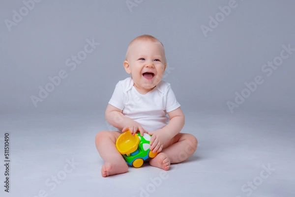 Fototapeta baby boy in a white bodysuit is sitting playing  with toy cars on white background