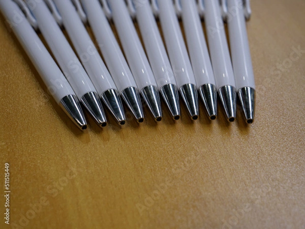Obraz Group of ten white plastic with metal tips ballpoint pens lying flat on beige lacquered wooden table top.
