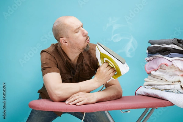 Fototapeta A funny bald man with emotions using electrical iron on ironing board over on blue background.