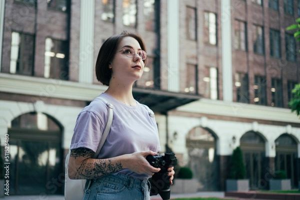 Fototapeta Portrait of Young Pretty Woman with Camera in Her Hands against Background of Modern Office Building