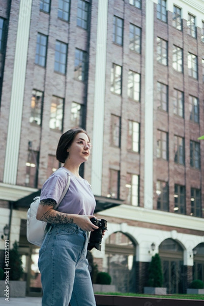 Fototapeta Portrait of Young Pretty Woman with Camera in Her Hands against Background of Modern Office Building