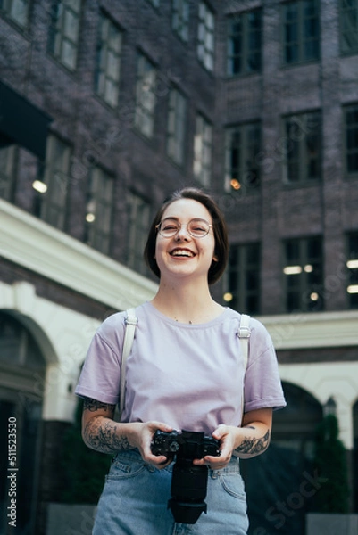 Fototapeta Portrait of Smiling Young Woman with Camera in Her Hands against Background of Modern Office Building