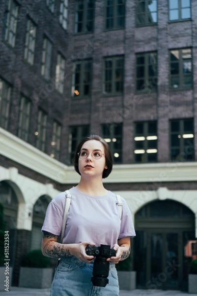 Fototapeta Portrait of Young Pretty Woman with Camera in Her Hands against Background of Modern Office Building