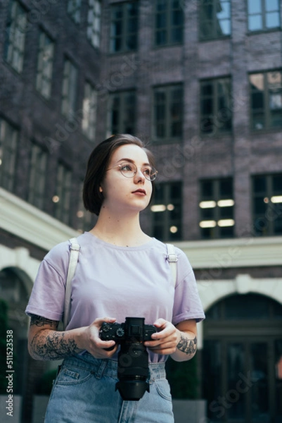Fototapeta Portrait of Young Pretty Woman with Camera in Her Hands against Background of Modern Office Building