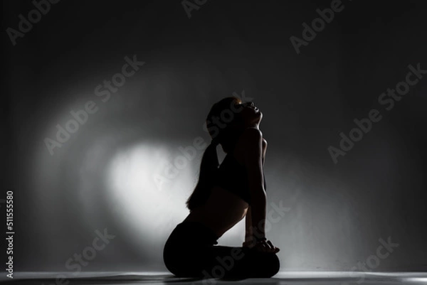 Fototapeta Fit girl resting and stretching after practicing yoga poses. Side lit half silhouette against dark background. .