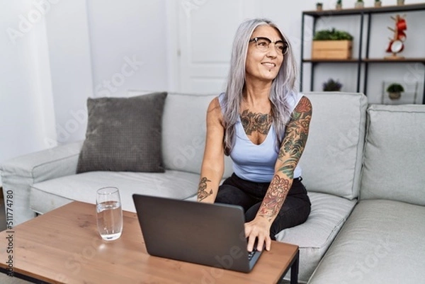 Fototapeta Middle age grey-haired woman using laptop at home looking away to side with smile on face, natural expression. laughing confident.