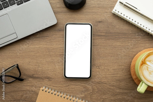 Fototapeta Smartphone with blank empty mockup screen is on top of wood office desk table with laptop computer and supplies. Top view with copy space, flat lay.