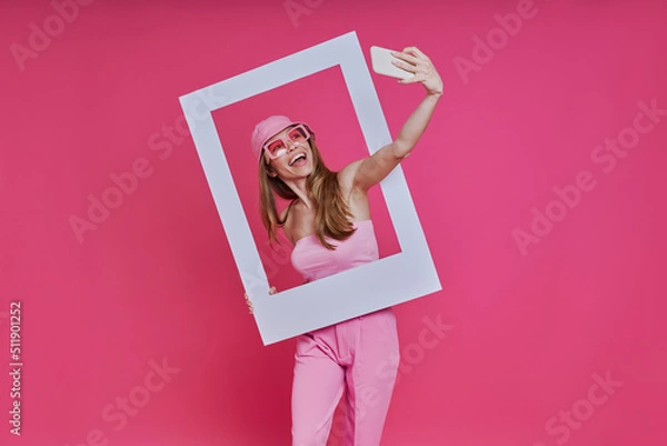 Fototapeta Playful young woman making selfie and looking through a picture frame while standing against pink background