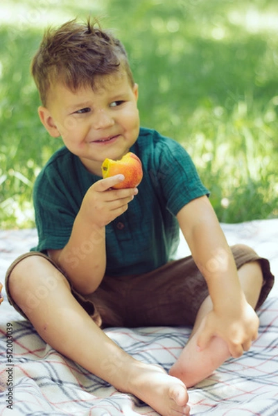 Fototapeta little brunettebeautiful boy eating a peach in summer park