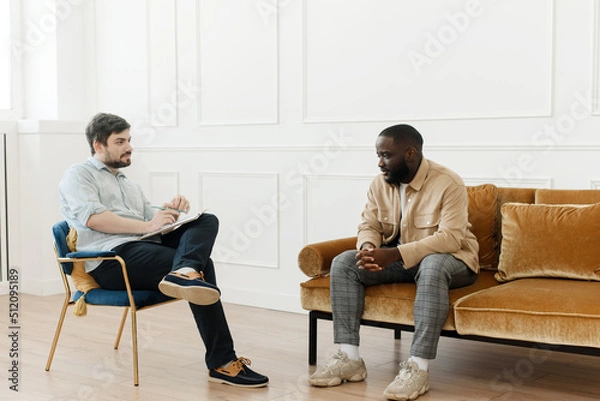 Fototapeta The therapist takes notes while talking to a depressed male patient and talks to an African American patient. Desperate black man suffering from nervous breakdown or depression having a session with a