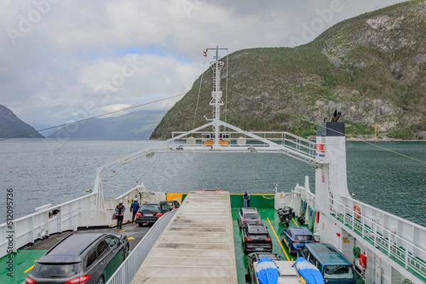 Fototapeta View of a typical Norwegian fjord landscape from a ferry upper deck. Blue sea water, cloudy sky and misty mountains. vehicles on the deck