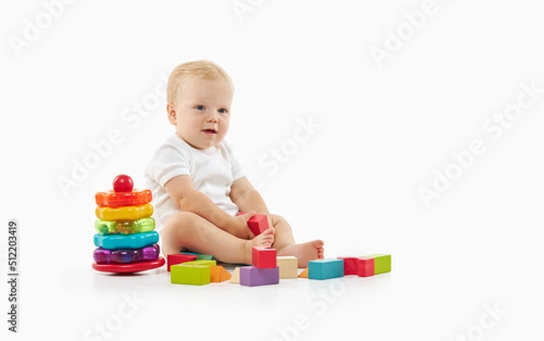 Fototapeta Cute baby plays with toys on a white isolated background. The child folds a constructor and a pyramid
