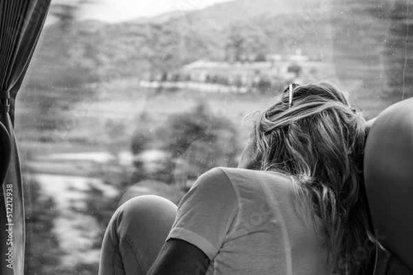 Fototapeta A black and white picture of a caucasian woman with long blond hair sitting in a bus staring outside the window of the landscape passing by with her raised knees on the bench.