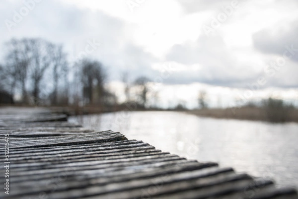 Fototapeta A dreamy picture of a landscape with a close up of an old wooden bridge over a lake with clouds and sunlight and trees in the background