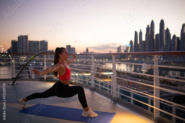 Obraz Fit woman does stretching in deep lunge on a on the outdoor in summer, , legs work out and hip joint mobility, enjoying morning aerobic classes with beautiful panoramic view of city soft focus
