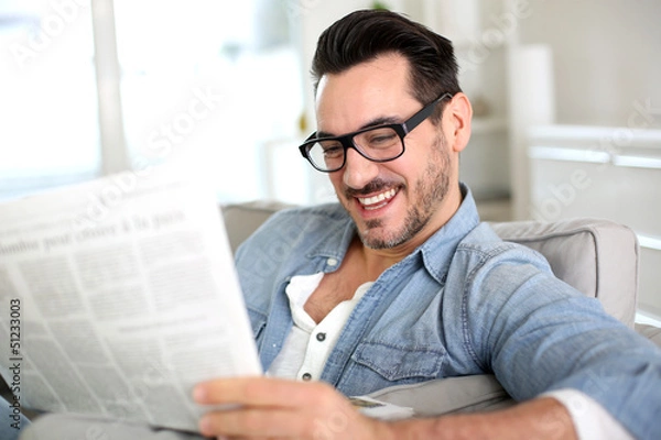 Fototapeta Cheerful guy reading newspaper in sofa