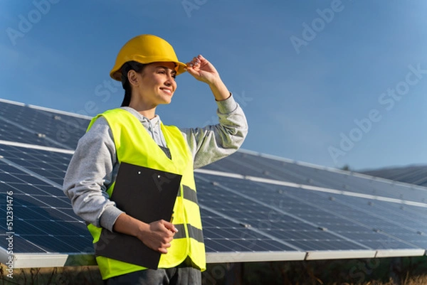 Fototapeta Professional woman engineer looking away with pleasure smile while working at ecological green field with solar panels. Photovoltaic power station concept
