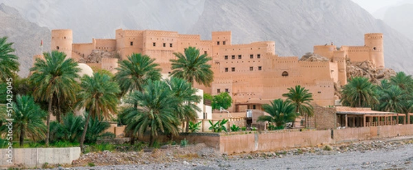 Fototapeta Nakhal, Oman - 04.02.2018: Panoramic shot of medieval Arabian military fortress. Very hot day with heat haze in the sky. Old Arabian castle. Tall stone walls. Palm trees in the front.