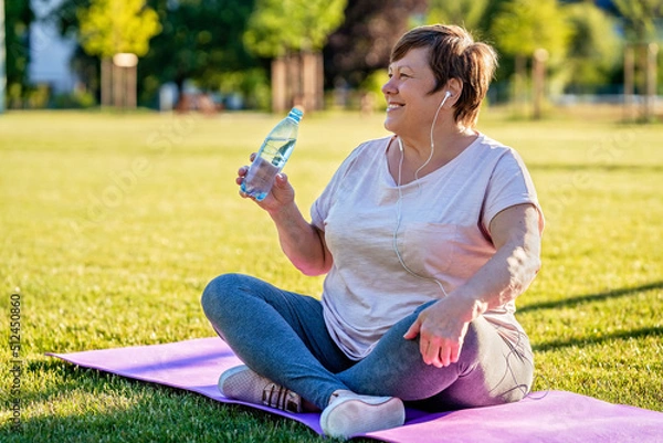 Fototapeta Relaxed senior plus size woman with earphones sitting on yoga mat on green grass outdoors resting after exercises drinking water at warm sunny summer day