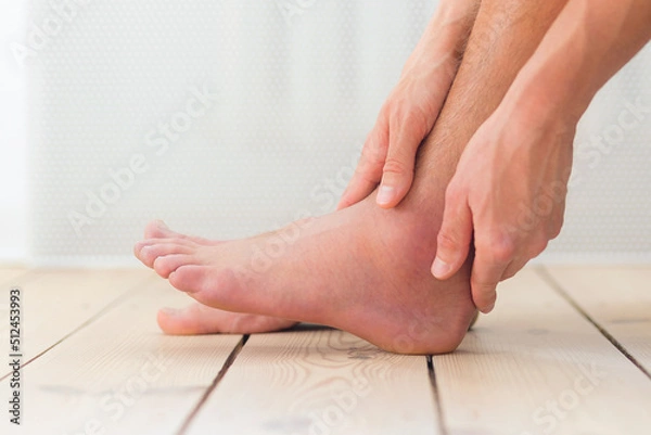 Obraz Sprained and swollen ankle. Man hold his sprained ankle sitting on the sofa at home. Ankle sprain. Injury.	