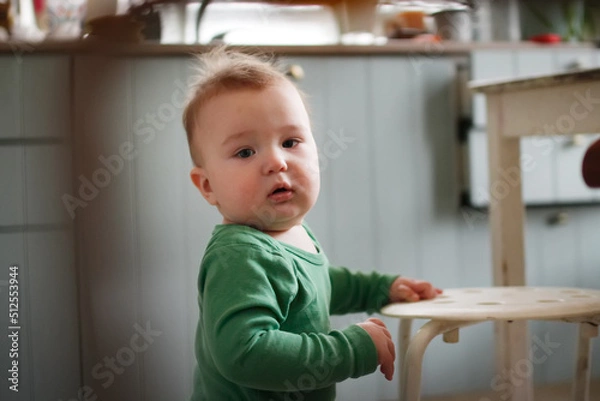 Fototapeta Funny one-year-old toddler standing at stool in kitchen, real people in real interior