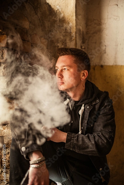 Fototapeta Portrait of a brutal smoking macho man in a black denim jacket with a stylish hairstyle, piercing in the eyebrow and lip and an earring in the ear. A rock musician or punk in an abandoned building
