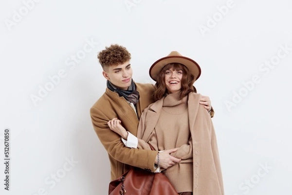 Fototapeta a beautiful, loving couple of a man and a woman stand on a white background in stylish autumn clothes, gently embracing and the woman happily looks at the camera, and the man at her