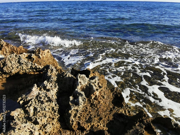 Fototapeta Long-hardened lava, porous, sharp coast of the Mediterranean Sea with oncoming waves against a sky with clouds.