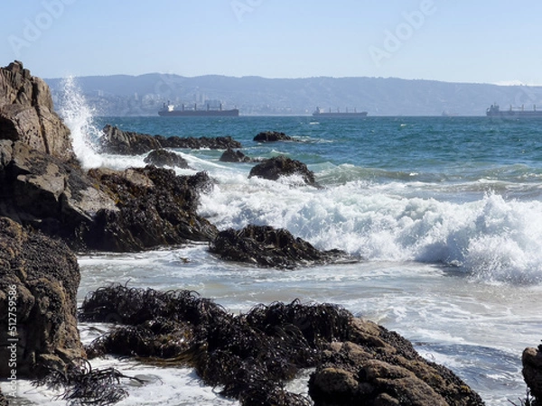 Fototapeta Sea water splash on rocks, algae and cargo ships by Valparaiso harbor on background. Brave waves crashing on shore on sunny day in Chile