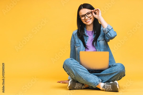 Fototapeta Business concept. Portrait of happy young woman in casual sitting on floor in lotus pose and holding laptop isolated over yellow background.