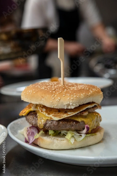 Fototapeta Restaurant kitchen imagery with a freshly prepared cheeseburger with toasted sesame bun, fresh vegetables, beef burger, melted cheese and fried egg resting on a plate and a busy chef in the background
