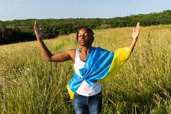 Fototapeta African american woman wrapped in ukrainian yellow blue flag flutters waving in the wind. National symbol of Ukraine. Stand with Ukraine, international support and solidarity
