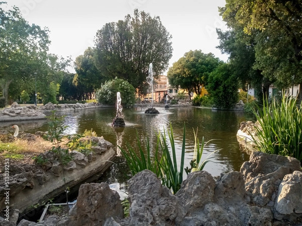 Fototapeta pleasant view of a fountain with 3 water jets in the pond of a park and panorama of the surrounding trees in a peaceful and natural evening atmosphere