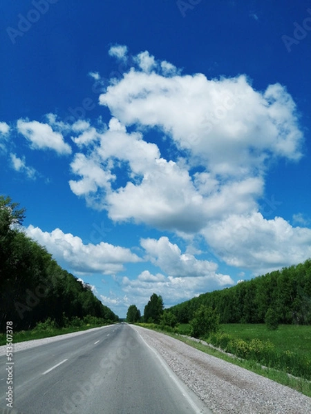 Fototapeta white clouds in the vast blue sky, road ahead with greenery on the sides, summer travel