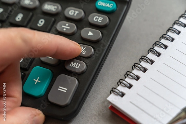 Fototapeta Close-up of male hand doing calculations on calculator. Index finger presses the percentage button. An open spring-loaded notebook and a pencil lie side by side on a gray surface. Selective focus.