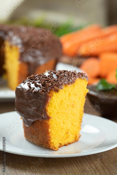 Fototapeta Close up piece of Brazilian carrot cake with chocolate frosting on wooden table with carrots in the background