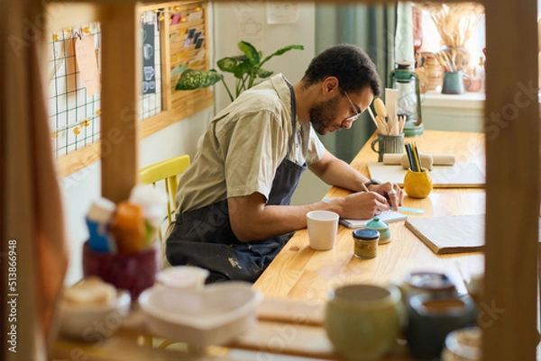 Fototapeta Young creative man bending over workplace while making sketch in notepad in spacious studio or workshop with displays for new items