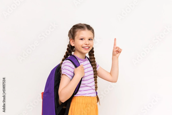 Obraz A child with a school backpack points up at your advertisement on a white isolated background. A little girl with pigtails points to the announcement of courses for children.