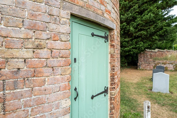 Fototapeta Exterior view of a very old round house, once a dove house. Showing the newly painted wood door. Seen in a rural graveyard.